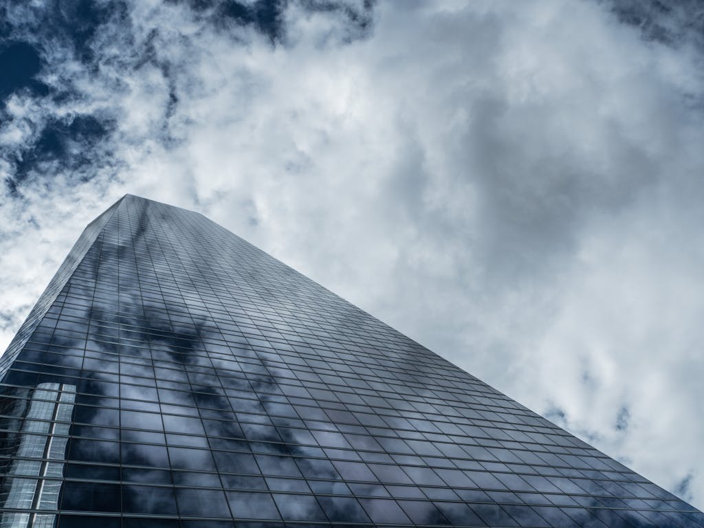 Low angle view of a towering modern skyscraper with glass windows reflecting clouds against a dramatic sky.