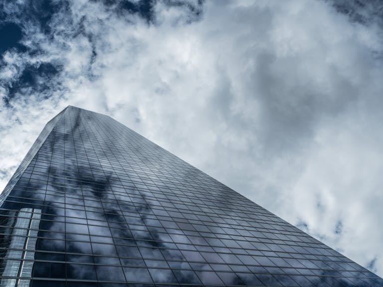 Low angle view of a towering modern skyscraper with glass windows reflecting clouds against a dramatic sky.